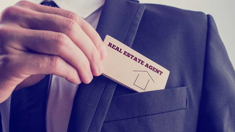 A real estate agent and homeowner discussing home documents at a kitchen table with a notepad and laptop.